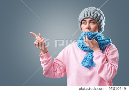 Young caucasian woman wearing knitted hat and scarf and point hand at left side. Gray background. The concept of energy crisis and winter season Young caucasian woman wearing knitted hat and scarf and point hand at left side. Gray background. The concept of energy crisis and winter season 95810649