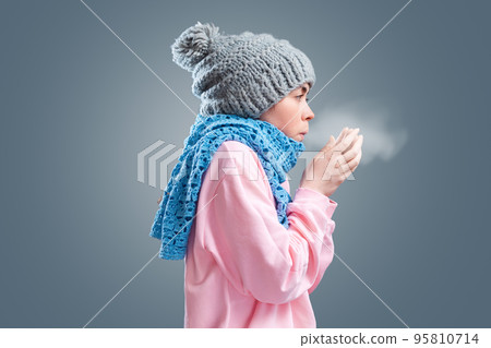 Portrait of young woman in a knitted hat and scarf is trying to keep warm, breathing to the hands. Gray background. The concept of energy crisis and winter season 95810714