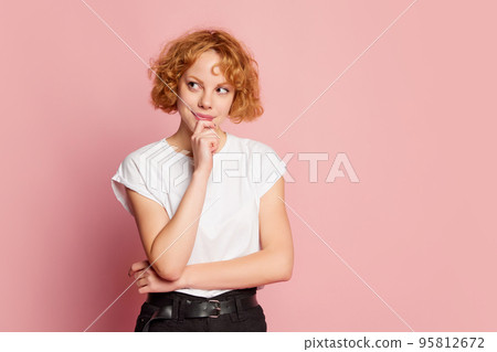 Half-length portrait of young red-haired girl, student thinking with light smile on her lips isolated on pink studio background. Fashion, facial expression, beauty concept. Half-length portrait of young red-haired girl, student thinking with light smile on her lips isolated on pink studio background. Fashion, facial expression, beauty concept. 95812672