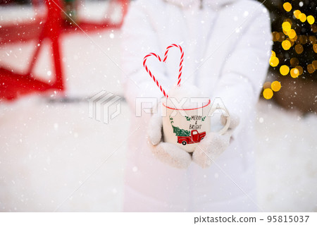 Woman with mug with snow, candy cane and inscription Merry and Bright in her hands outdoor in warm clothes in winter festive market. Fairy lights garlands decorated snow town for new year. Christmas Woman with mug with snow, candy cane and inscription Merry and Bright in her hands outdoor in warm clothes in winter festive market. Fairy lights garlands decorated snow town for new year. Christmas 95815037