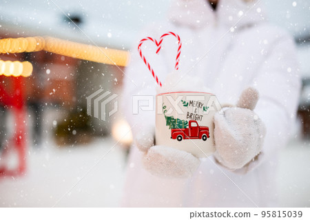 Woman with mug with snow, candy cane and inscription Merry and Bright in her hands outdoor in warm clothes in winter festive market. Fairy lights garlands decorated snow town for new year. Christmas Woman with mug with snow, candy cane and inscription Merry and Bright in her hands outdoor in warm clothes in winter festive market. Fairy lights garlands decorated snow town for new year. Christmas 95815039