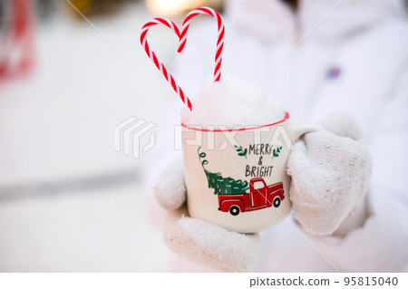 Woman with mug with snow, candy cane and inscription Merry and Bright in her hands outdoor in warm clothes in winter festive market. Fairy lights garlands decorated snow town for new year. Christmas Woman with mug with snow, candy cane and inscription Merry and Bright in her hands outdoor in warm clothes in winter festive market. Fairy lights garlands decorated snow town for new year. Christmas 95815040