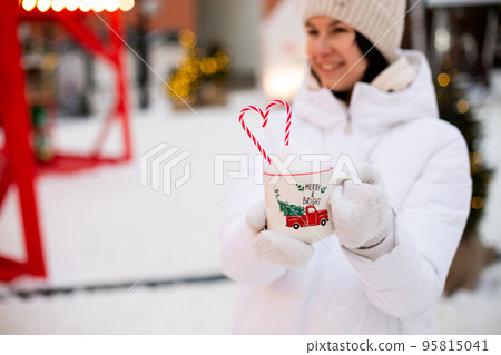 Woman with mug with snow, candy cane and inscription Merry and Bright in her hands outdoor in warm clothes in winter festive market. Fairy lights garlands decorated snow town for new year. Christmas Woman with mug with snow, candy cane and inscription Merry and Bright in her hands outdoor in warm clothes in winter festive market. Fairy lights garlands decorated snow town for new year. Christmas 95815041