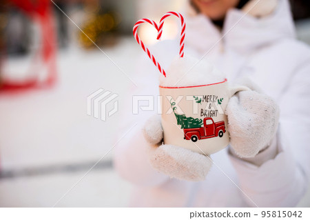 Woman with mug with snow, candy cane and inscription Merry and Bright in her hands outdoor in warm clothes in winter festive market. Fairy lights garlands decorated snow town for new year. Christmas Woman with mug with snow, candy cane and inscription Merry and Bright in her hands outdoor in warm clothes in winter festive market. Fairy lights garlands decorated snow town for new year. Christmas 95815042