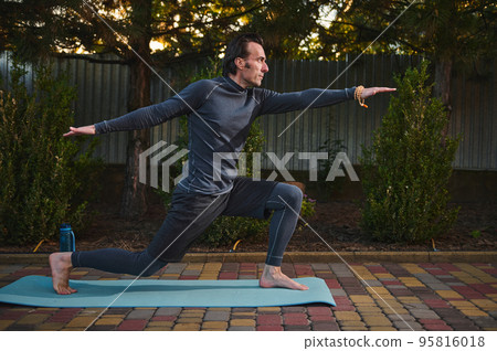 Side full-length portrait of a middle-aged Caucasian active man, an athlete exercising outdoors, practicing yoga at sunset, assuming warrior pose, standing barefoot on a fitness mat. Practice. Asana. Side full-length portrait of a middle-aged Caucasian active man, an athlete exercising outdoors, practicing yoga at sunset, assuming warrior pose, standing barefoot on a fitness mat. Practice. Asana. 95816018