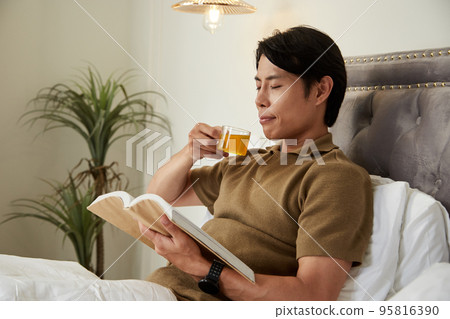 Happy calm young man drinking hebal tea and reading good book in bed 95816390