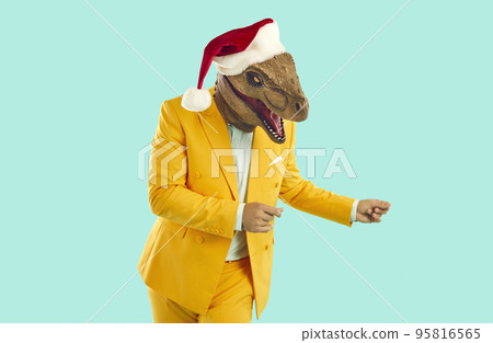 Funny showman in yellow suit, Santa hat and rubber dinosaur mask dancing on turquoise background. 95816565