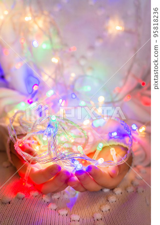 Christmas garland of multi-colored led-bulbs in the hands of a child dressed in a white knitted sweater. The concept of comfort, warmth, the advent of Christmas Christmas garland of multi-colored led-bulbs in the hands of a child dressed in a white knitted sweater. The concept of comfort, warmth, the advent of Christmas 95818236