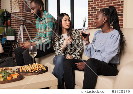 Adult asian female listening, hearing attentively african american partner conversation, discussion, talk at party.Diverse friends drinking wine, eating bread sticks, cheese at a colleagues meeting. Adult asian female listening, hearing attentively african american partner conversation, discussion, talk at party.Diverse friends drinking wine, eating bread sticks, cheese at a colleagues meeting. 95820179