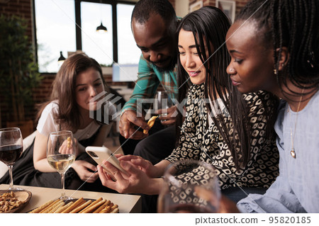 Cultural diverse mates, friends, pointing at phone display, smiling, talking, discussing at house, home living room multiethnic gathering. Various ethnicities people watching movies on smartphone. Cultural diverse mates, friends, pointing at phone display, smiling, talking, discussing at house, home living room multiethnic gathering. Various ethnicities people watching movies on smartphone. 95820185