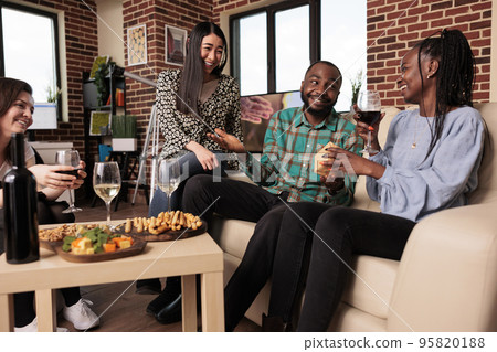 Varying ethnicities people group exchanging gifts at birthday party. African american woman being surprised with present from friends at gathering, drinking wine enjoying snacks. Varying ethnicities people group exchanging gifts at birthday party. African american woman being surprised with present from friends at gathering, drinking wine enjoying snacks. 95820188