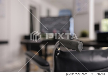 Close up view of wheelchair in disabled friendly office. In the background unfocused view of a tabletop computer with yearly charts results in financial area, interior workplace. Close up view of wheelchair in disabled friendly office. In the background unfocused view of a tabletop computer with yearly charts results in financial area, interior workplace. 95820281