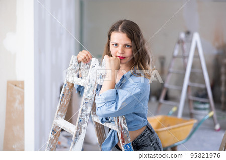 Attractive young woman in denim shirt and shorts posing next to stepladder in refurbished room 95821976