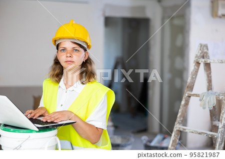 Female foreman in a protective helmet and a yellow vest checks the execution of repair work using laptop 95821987
