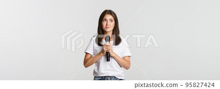 Shy cute brunette girl scared of singing in public, standing with microphone and looking nervous 95827424