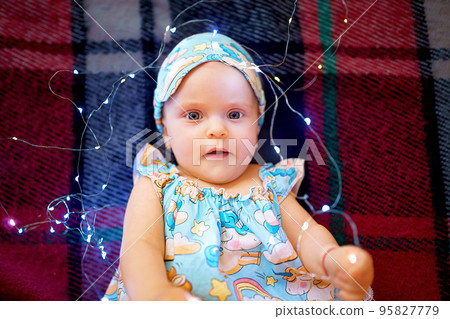 Top view of cute baby girl in pajamas playing with christmas lights while lying on checkered plaid  95827779