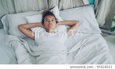 Sad race lady is lying in bed and sighing looking at ceiling then closing her eyes and falling asleep with smile. Comfortable bed, soft pillows and sheets are visible. 95828021