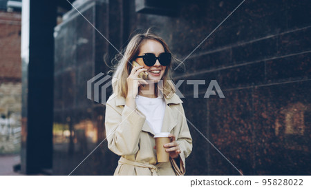 Beautiful young woman with blond hair is talking on mobile phone and drinking take away coffee walking in modern city along street and enjoying to-go drink and conversation. 95828022