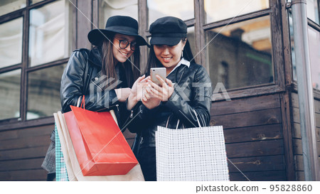 Excited girls friends are using smartphone touching screen standing in the street and holding shopping bags on autumn day. Modern technology and youth concept. 95828860