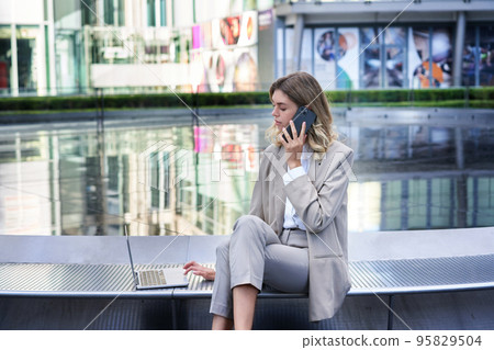 Portrait of busy woman sitting in city centre with laptop and mobile phone, corporate lady calling someone and checking work on computer Portrait of busy woman sitting in city centre with laptop and mobile phone, corporate lady calling someone and checking work on computer 95829504