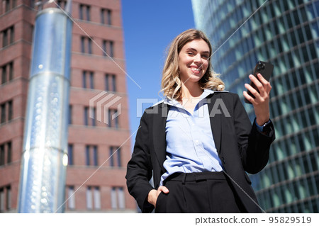 Low angle shot of businesswoman in suit, standing on street and looking at mobile phone, holding smartphone 95829519