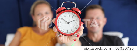 Smiling man and woman lying in bed and holding alarm clock at ten o'clock Smiling man and woman lying in bed and holding alarm clock at ten o'clock 95830786