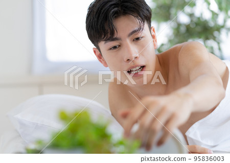 Young man waking up in bed in the morning and trying to turn off the alarm clock 95836003