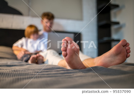 Ginger man and his cute son lying in bed with their gadgets 95837090