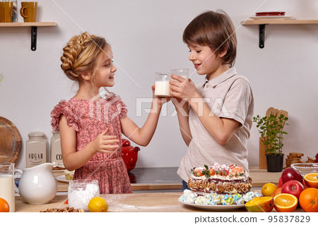Little friends are making a cake together at a kitchen against a white wall with shelves on it. Little friends are making a cake together at a kitchen against a white wall with shelves on it. 95837829