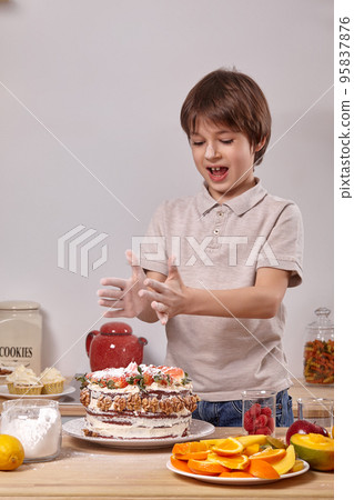 Little boy is making a homemade cake with an easy recipe at kitchen against a white wall with shelves on it. 95837876