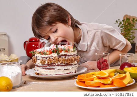 Little boy is making a homemade cake with an easy recipe at kitchen against a white wall with shelves on it. 95837881