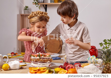 Little friends are making a cake together at a kitchen against a white wall with shelves on it. 95837923