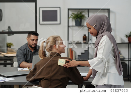 Muslim woman putting notepaper with word fool on back of blond businesswoman 95837933