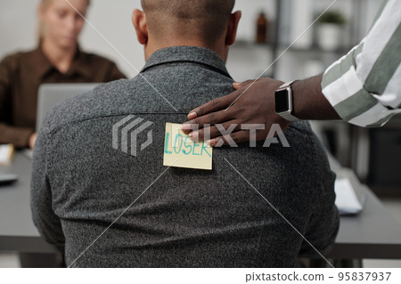 Hand of black man putting sticker with written word loser on back of colleague Hand of black man putting sticker with written word loser on back of colleague 95837937