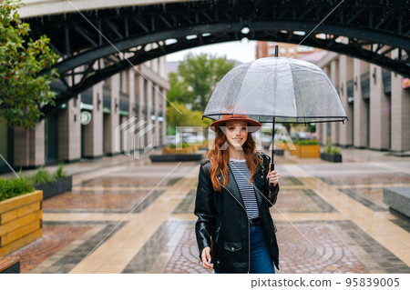 Front view of happy young woman wearing fashion hat walking on European city street with transparent umbrella enjoying rainy weather outdoors, smiling looking at camera. Concept of female lifestyle. 95839005