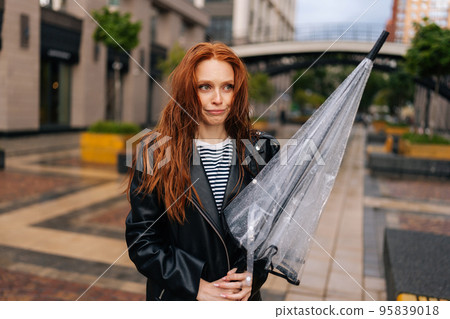Portrait of disgruntled attractive young woman with long red-hair standing with wet, disheveled hair after being caught in cold autumn rain, holding in hand broken umbrella, sad looking away. 95839018