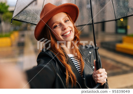 Smiling redhead young woman wearing elegant hat under transparent umbrella making selfie photo looking at camera, holding mobile phone on hand, standing in rain on European city street 95839049