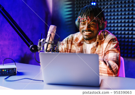 African radio host sitting at desk recording in studio with microphone and laptop 95839063
