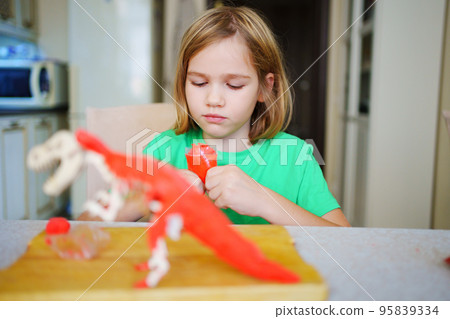 A blonde little girl makes a dinosaur craft out of wood and air plasticine.  95839334