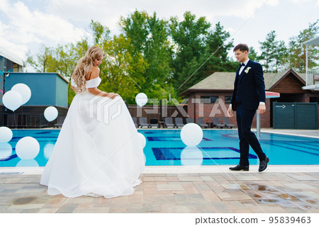 bride and groom in classic clothes by the pool decorated with white balloons. bride and groom in classic clothes by the pool decorated with white balloons. 95839463