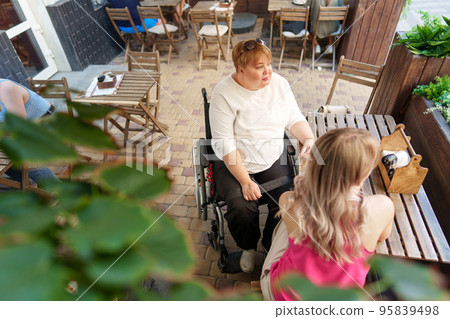 Mother with disability in wheelchair talking to her daughter while sitting at the table in cafe Mother with disability in wheelchair talking to her daughter while sitting at the table in cafe 95839498