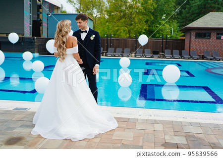 bride and groom in classic clothes by the pool decorated with white balloons. bride and groom in classic clothes by the pool decorated with white balloons. 95839566