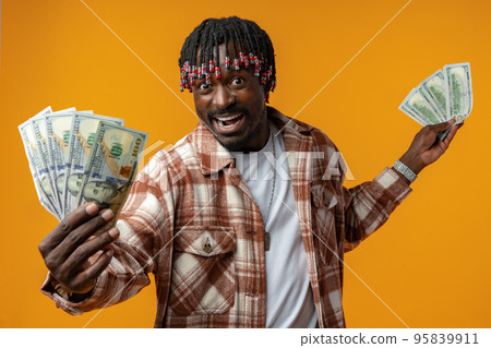 Young happy rich african man in casual shirt holding dollar bills against yellow background 95839911