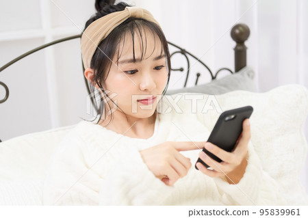 A young woman lying on the bed and operating a smartphone A young woman lying on the bed and operating a smartphone 95839961