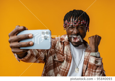 Young african man dressed in casual playing games on mobile phone over yellow background. 95840093
