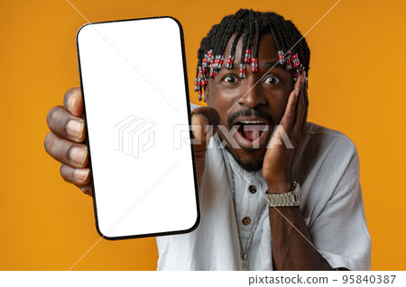 Excited young african man holding smartphone with white blank screen in hand on yellow studio background Excited young african man holding smartphone with white blank screen in hand on yellow studio background 95840387