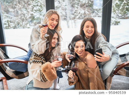 Young women enjoying winter weekends inside contemporary barn house. Four girls having fun, taking selfies on phone and drinking wine. Young women enjoying winter weekends inside contemporary barn house. Four girls having fun, taking selfies on phone and drinking wine. 95840539