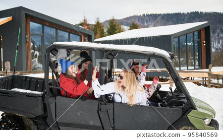 Happy women riding on off-road buggy car, giving high five to each other. Contemporary barnhouse on background. Concept of active leisure and winter activities. Happy women riding on off-road buggy car, giving high five to each other. Contemporary barnhouse on background. Concept of active leisure and winter activities. 95840569