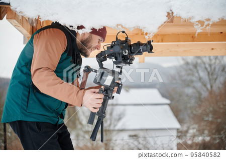 Happy videographer man shooting footage, using camera mounted on gimbal stabilizer equipment in winter. 95840582