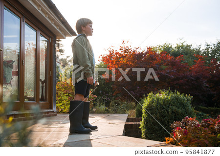 School kid boy in stylish rubber wellington boots ready for walk in autumn park. Gum boot rain boot. House in nice autumn garden. Happy child enjoy nature. 95841877
